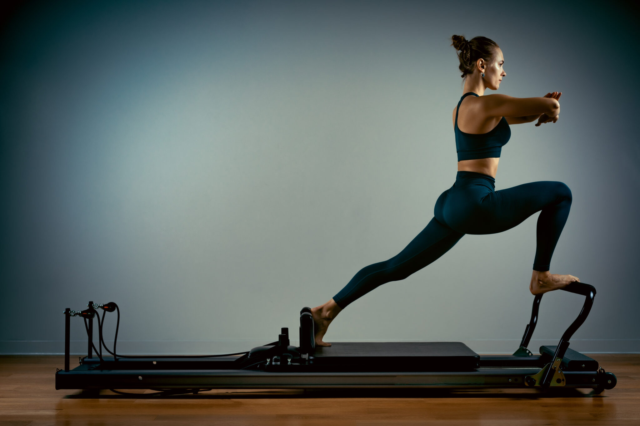 Young girl doing pilates exercises with a reformer bed. Beautiful slim fitness trainer on a reformer gray background, low key, art light, copy space advertising banner.